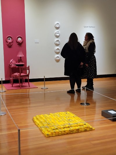 An art gallery. Closest to the viewer, there is an installation of yellow soap bars. On the wall, there are several round white artworks and a pink art installation. There are two figures are standing between the yellow and the pink installations talking.